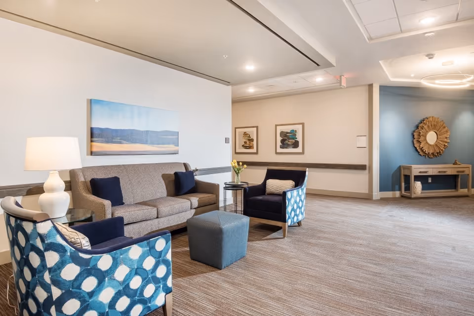 A cozy seating area in a senior living facility featuring a beige sofa with two dark blue pillows, two blue patterned armchairs, a small square ottoman, and a side table with a vase of yellow flowers. The walls are decorated with landscape paintings and abstract art, and there is a wooden console table with a decorative round mirror on a blue accent wall in the background.