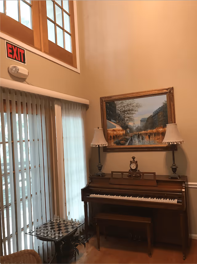 A cozy corner of a room featuring a wooden upright piano with a matching bench, flanked by two table lamps with fringed shades. Above the piano hangs a framed painting depicting a street scene with the Eiffel Tower in the background. To the left, there are vertical blinds covering a large window or sliding door, with an exit sign above. A small table with a chessboard and pieces is positioned near the window.