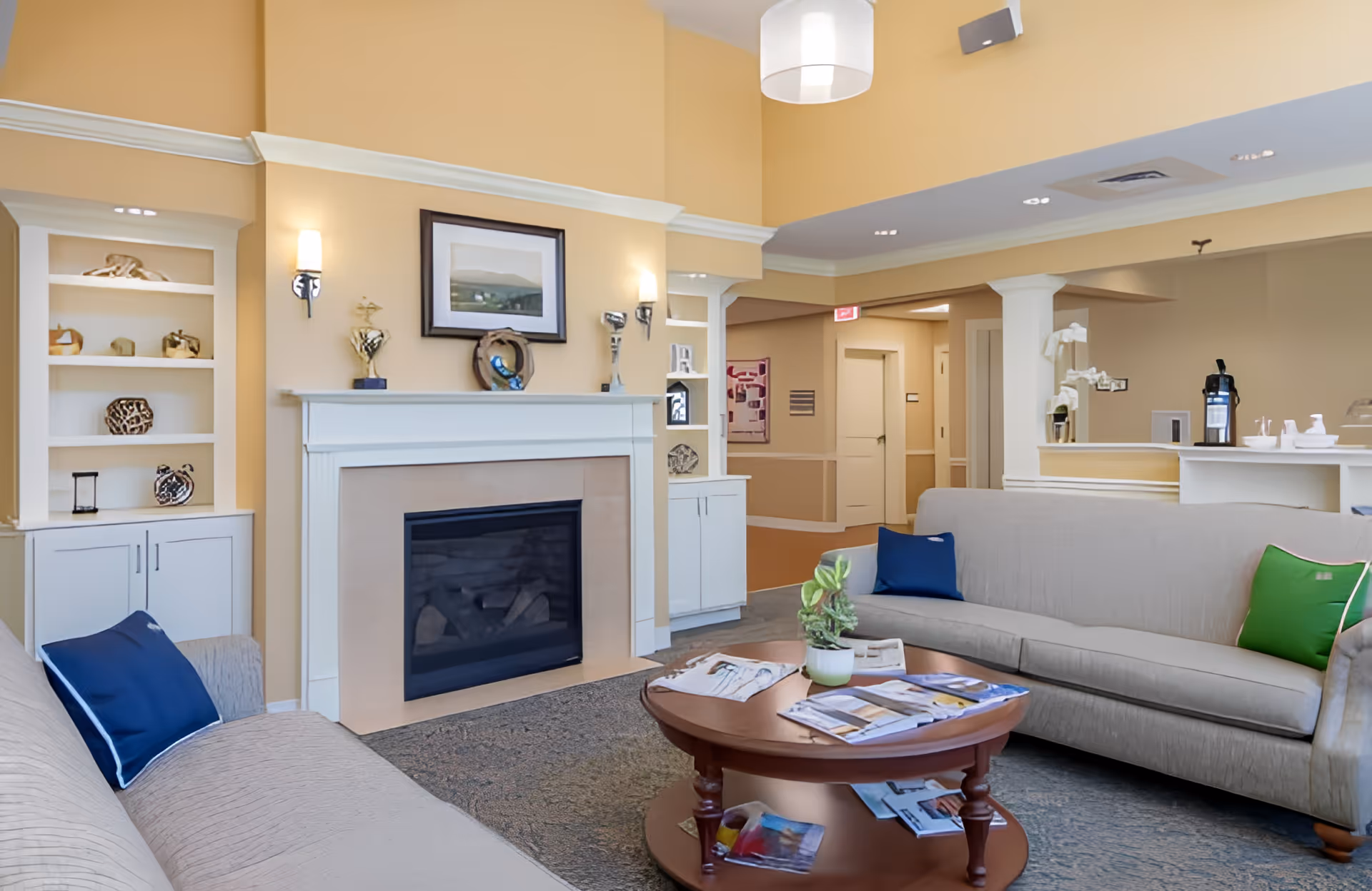 A cozy living room area with two beige sofas adorned with blue and green pillows, a wooden coffee table with magazines and a small plant, a fireplace with decorative items on the mantel, built-in shelves with ornaments, and warm yellow walls.