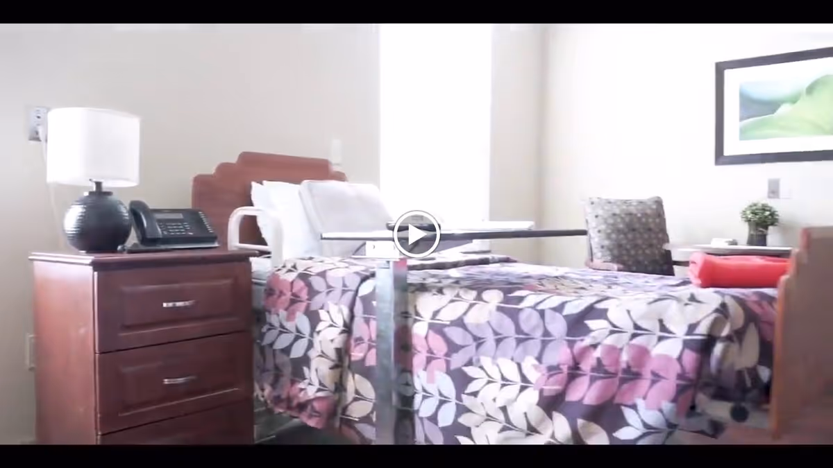 A furnished senior bedroom with a patterned bed, bedside table holding a lamp and phone, an overbed table, and a chair.