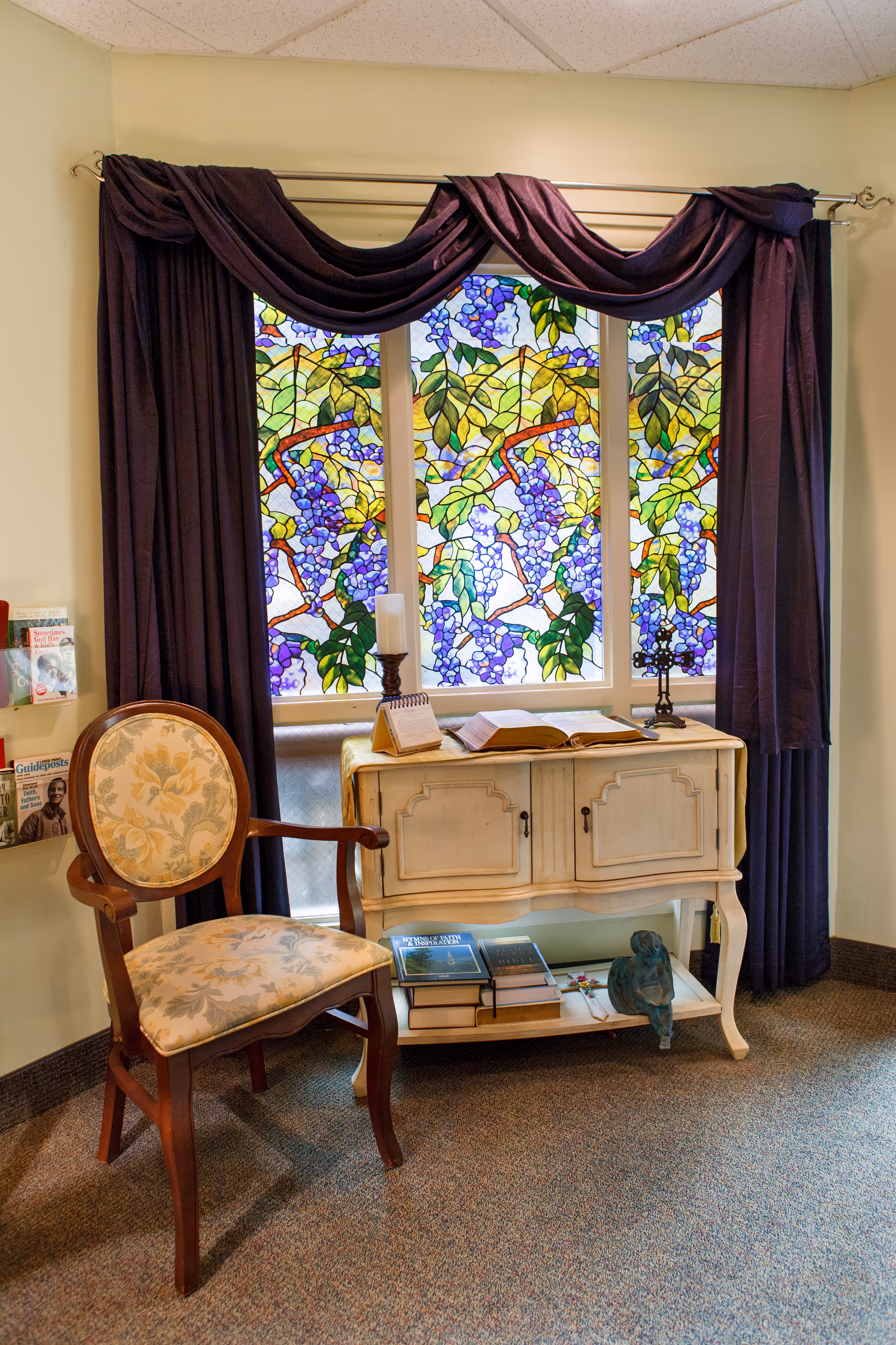 A cozy interior seating area with a floral upholstered chair, a small cream cabinet holding books and candles, and a colorful stained-glass window framed by dark drapes.