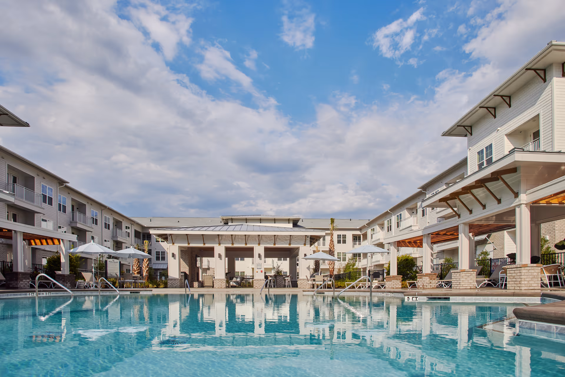 A large outdoor swimming pool and courtyard surrounded by multi-story senior living buildings with umbrellas and lounge chairs under a partly cloudy sky.