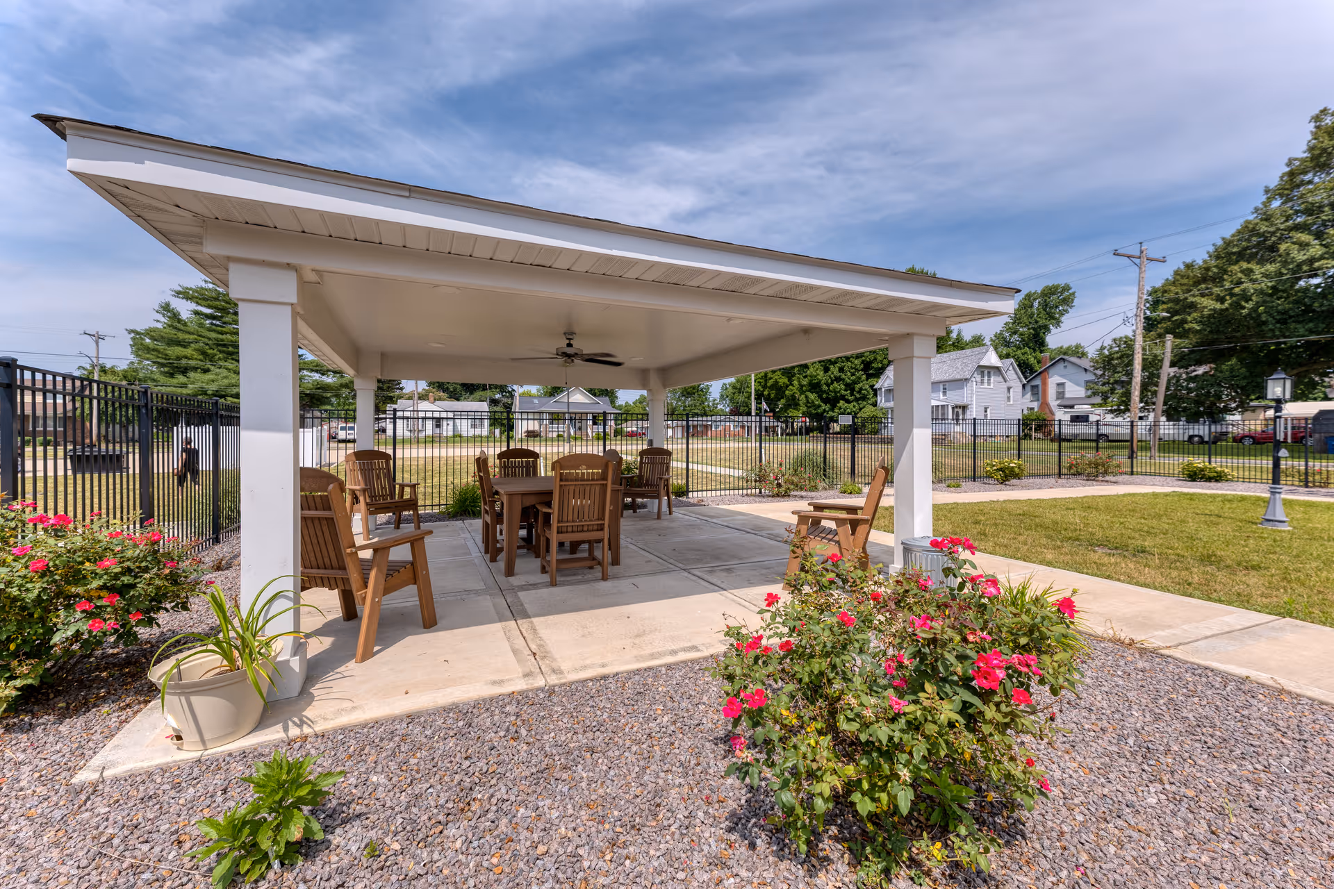Outdoor covered seating area with wooden chairs and tables under a white pavilion roof, surrounded by a garden with blooming red flowers and a fenced grassy yard in the background under a partly cloudy sky.