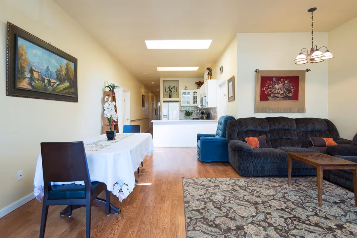 A bright and cozy living and dining area with wooden flooring. The dining area has a table covered with a white tablecloth and two dark wooden chairs. The living area features a dark blue sectional sofa with orange and purple cushions, a wooden coffee table, and a patterned rug. The walls are decorated with a landscape painting and a floral tapestry. The kitchen is visible in the background with white cabinets and a refrigerator. Two skylights provide natural light to the space.