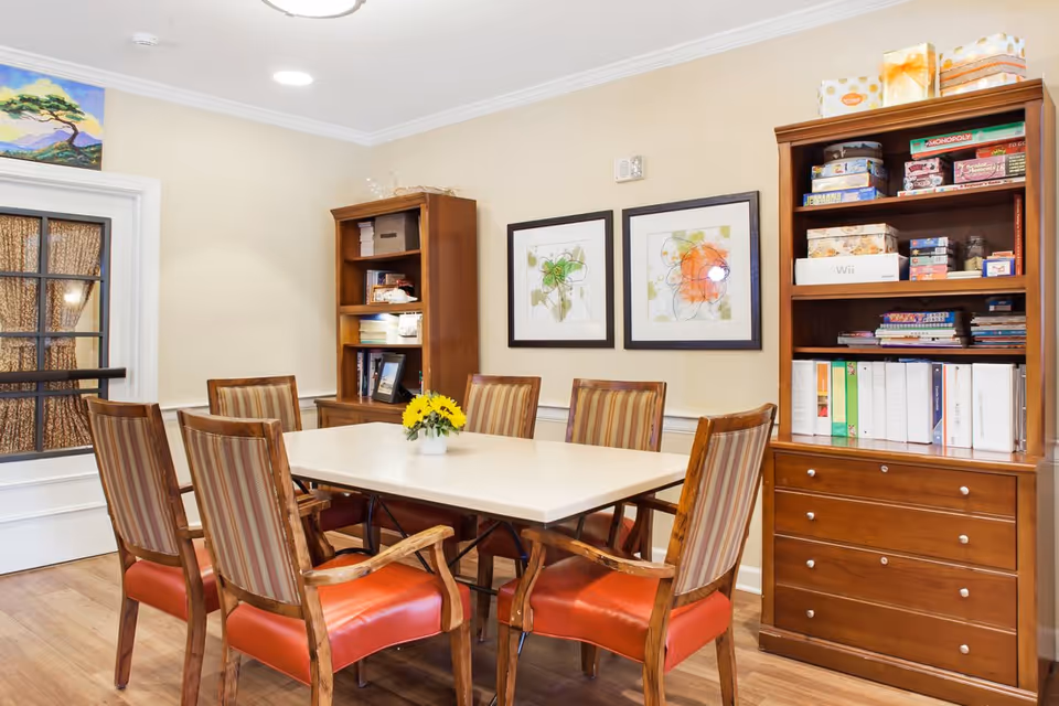 A well-lit room with a rectangular white table surrounded by six wooden chairs with red cushioned seats and striped backs. Two wooden bookshelves filled with books, board games, and decorative items are against the beige walls. Two framed abstract paintings hang on the wall above the table. A small vase with yellow flowers is centered on the table. A door with glass panels and curtains is visible on the left side.
