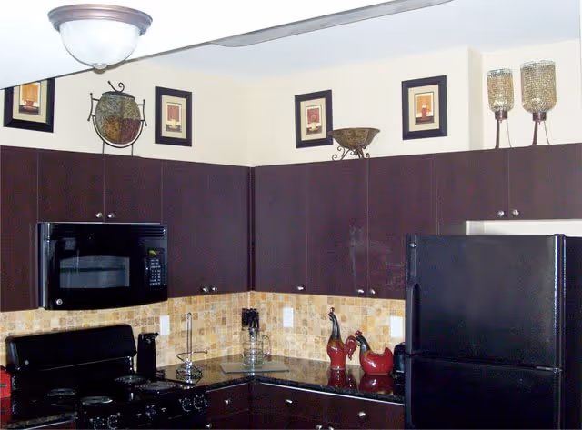 Modern kitchen with dark wood cabinets, black appliances including a microwave, stove, and refrigerator. The backsplash is made of beige and brown tiles. Decorative items such as framed pictures, a bowl, and candle holders are placed on top of the cabinets.