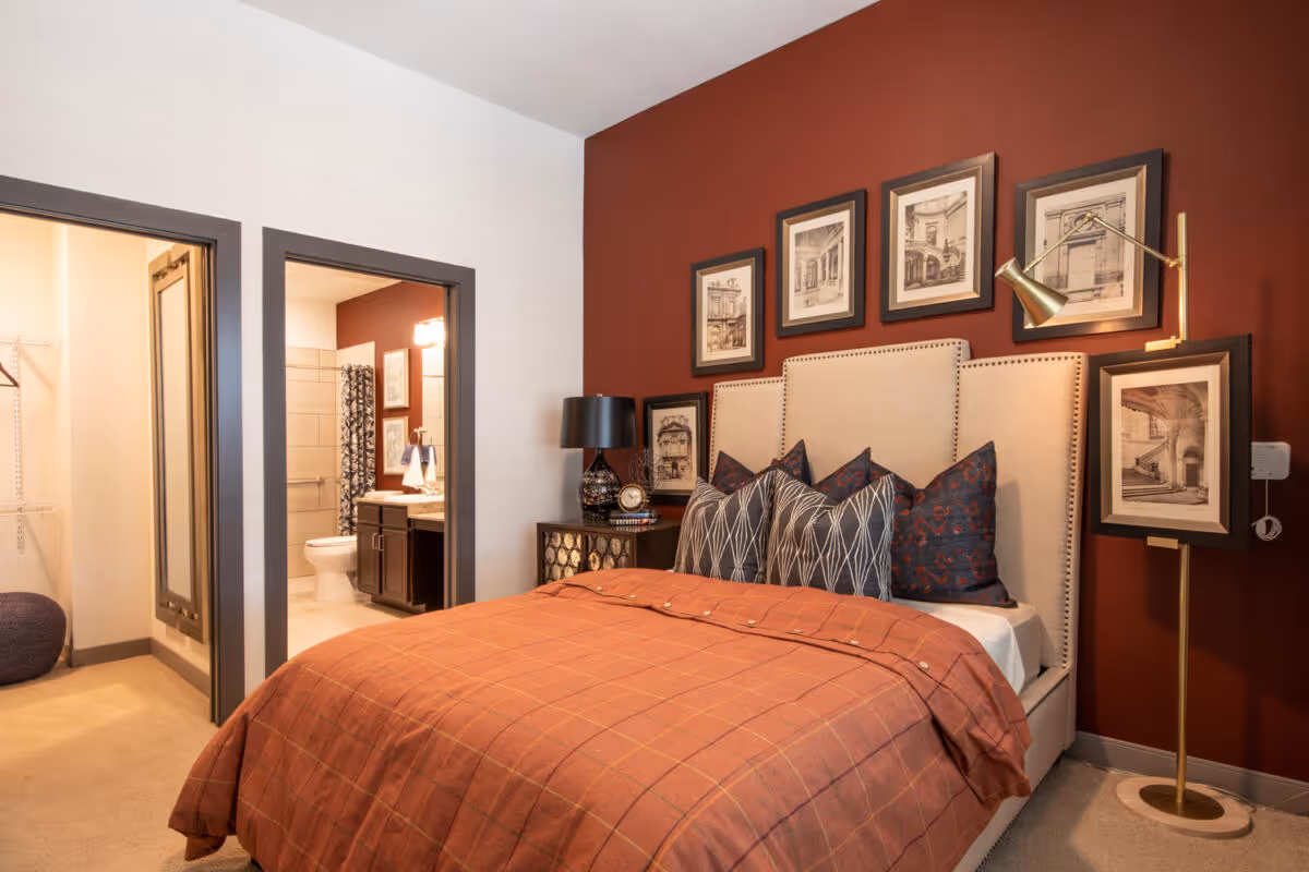 A cozy bedroom with a beige upholstered bed featuring multiple patterned pillows and an orange plaid blanket. The wall behind the bed is painted dark red and decorated with six framed architectural sketches. To the right of the bed is a gold floor lamp holding one of the framed pictures. To the left is a nightstand with a black lamp and a clock. Two doorways are visible: one leading to a walk-in closet and the other to a bathroom with a toilet, sink, and shower curtain.