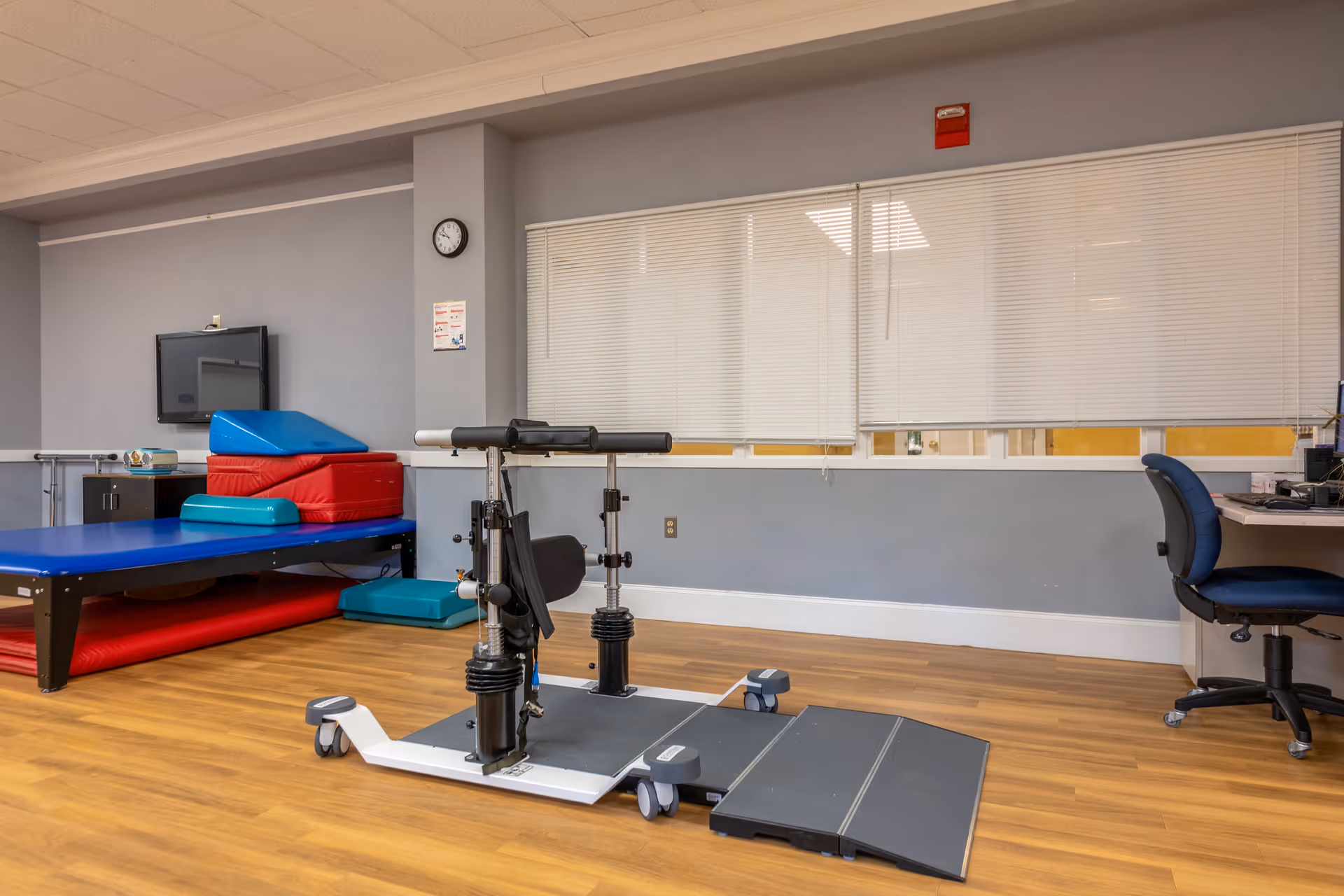 A physical therapy or rehabilitation room with padded exercise mats stacked in the corner, a blue therapy table, a piece of exercise equipment with adjustable supports on wheels, a desk with a blue office chair, and a wall-mounted TV. The room has wooden flooring and a large window with closed blinds.