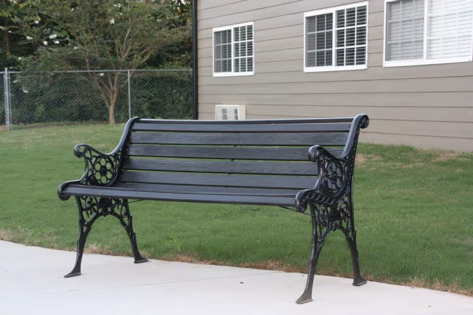 A black metal and wood outdoor bench placed on a concrete sidewalk with green grass and a building with windows in the background.