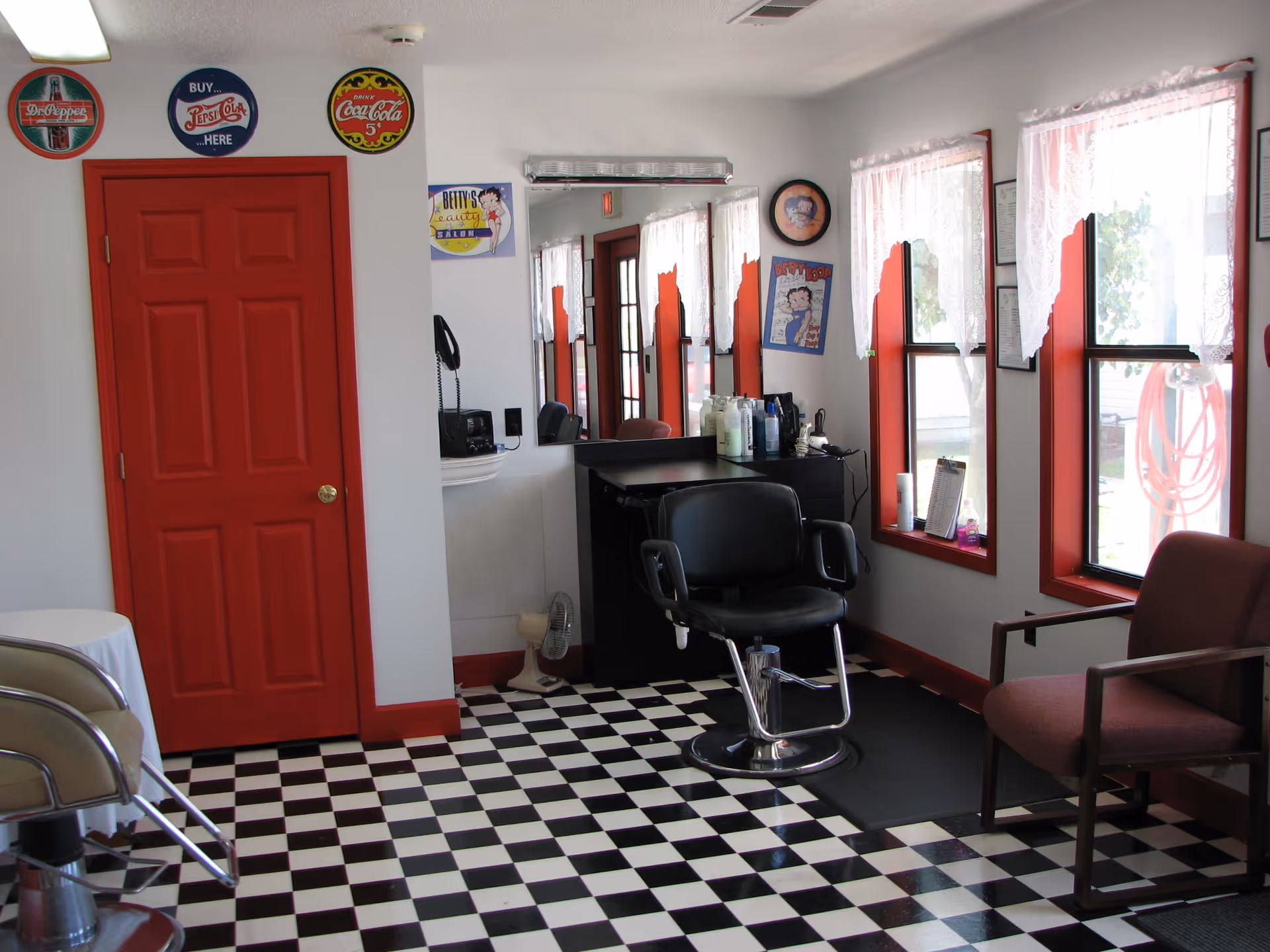Interior of a retro-style salon with a black barber chair, checkerboard floor, red door and window trim, and a mirror with hair products.