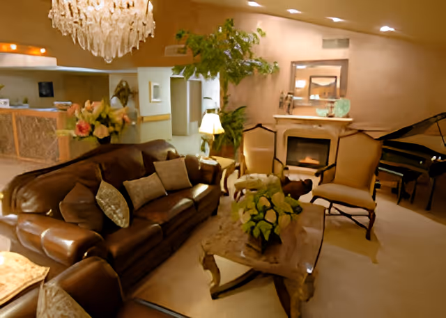 A cozy living room area in a senior care facility featuring a brown leather sofa with several cushions, two armchairs, a wooden coffee table with a floral arrangement, a fireplace with a mirror above it, a grand piano in the corner, a chandelier, and various plants and lamps providing warm lighting.