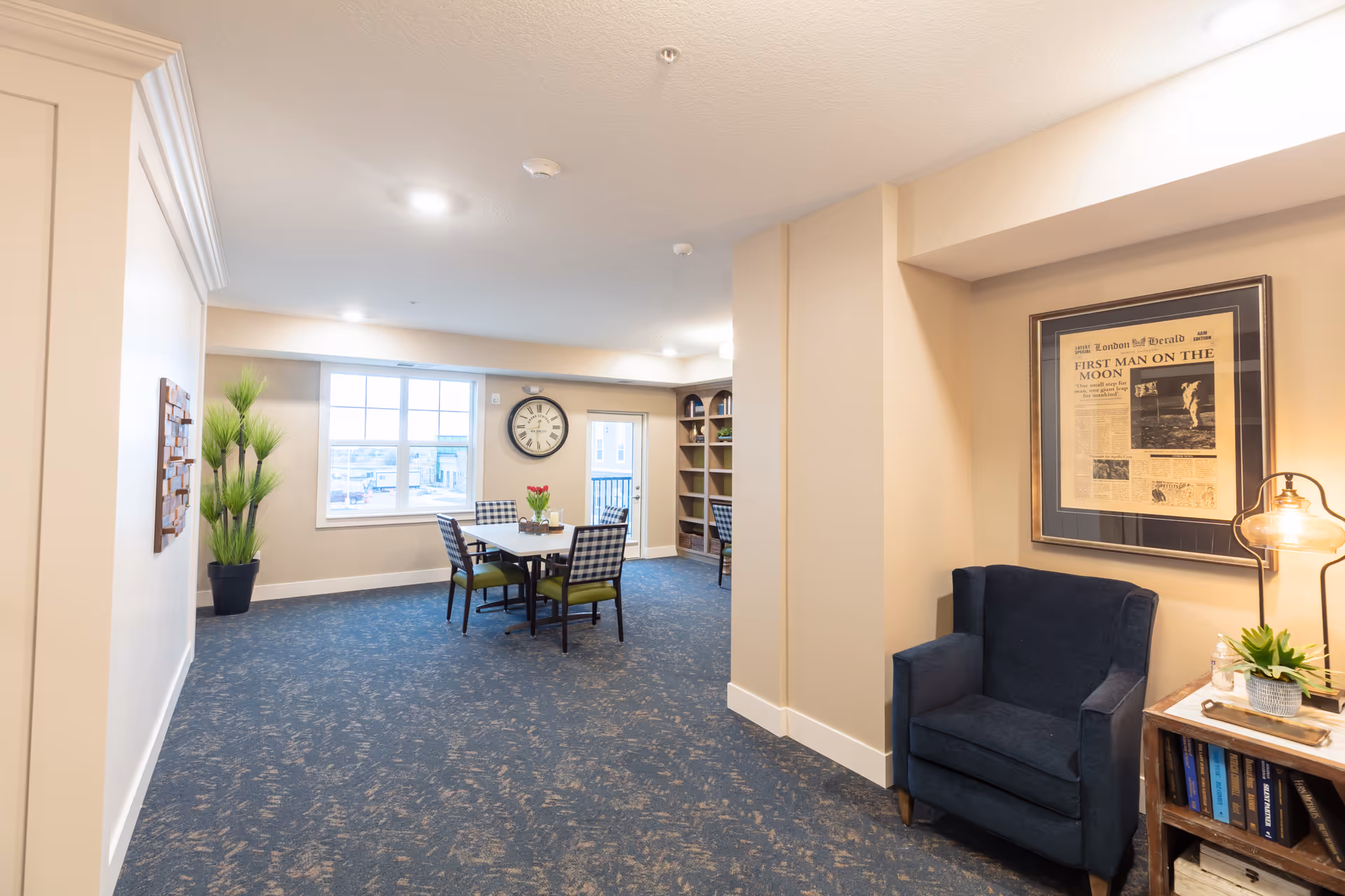 A bright communal lounge and dining area with a table and chairs, a blue armchair, bookshelf, and wall clock.
