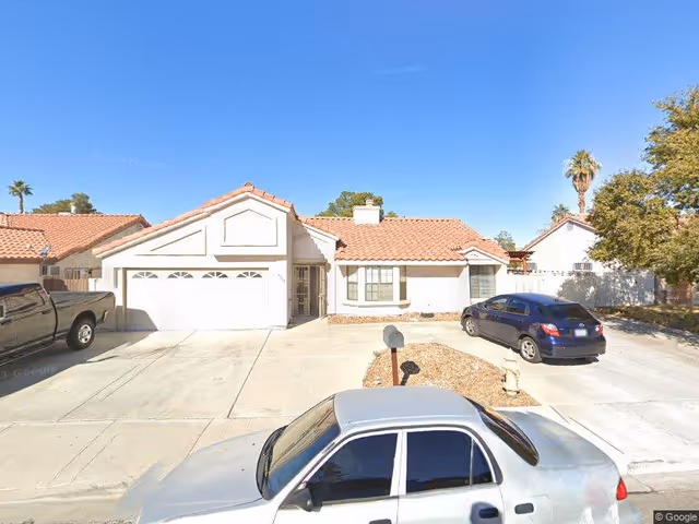 Front exterior view of a single-story residential building with a tiled roof, a two-car garage, and a driveway with three parked vehicles under a clear blue sky.