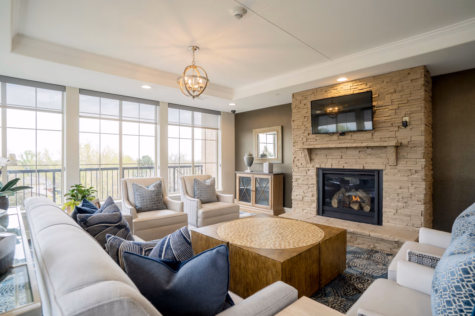 A bright and cozy living room with large windows letting in natural light. The room features a stone fireplace with a mounted flat-screen TV above it, a round wooden coffee table, a beige sofa with multiple blue and patterned pillows, and two beige armchairs with patterned cushions. There is a sideboard with decorative items and a mirror on the wall behind the armchairs.