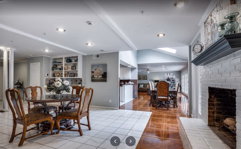 Interior view of a senior living facility showing a dining area with a round glass table and four wooden chairs with cushions on a white tiled floor. Adjacent to this area is a larger dining space with a long wooden table and multiple chairs on a polished wooden floor. The room features a white brick fireplace on the right, built-in shelves with decorative items on the left, and a ceiling with recessed lighting.