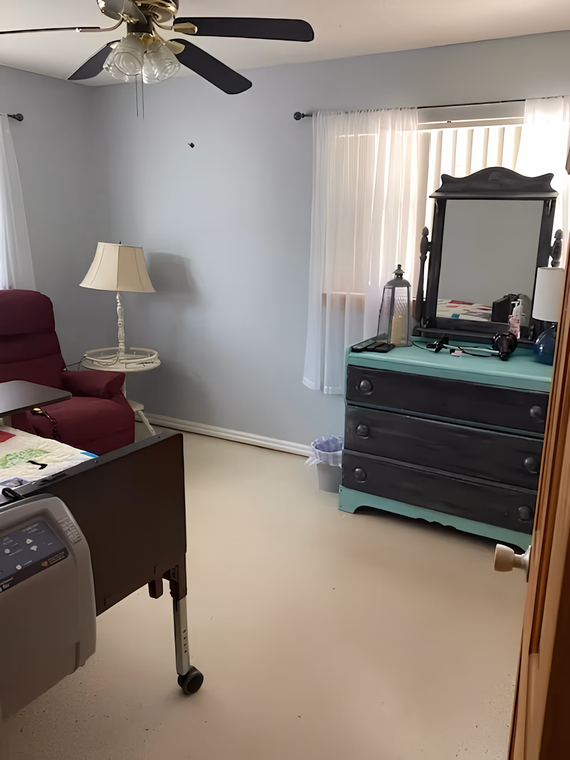 A small bedroom in an assisted living facility with a hospital-style bed, a maroon recliner chair, a white side table with a lamp, a ceiling fan with lights, and a black and turquoise dresser with a mirror. The room has light-colored walls and a window with sheer white curtains.