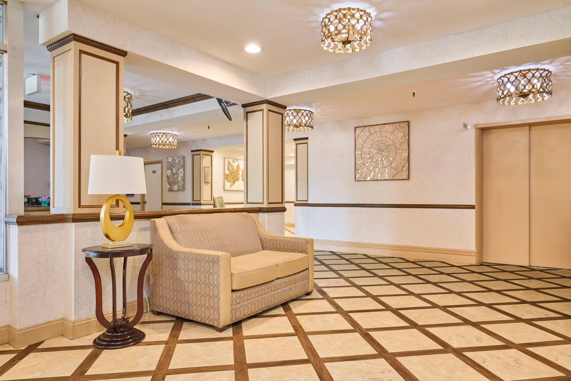 A well-lit interior common area with a patterned tile floor, beige walls, and decorative ceiling lights. There is a beige patterned loveseat next to a small round wooden table with a modern lamp on it. The area includes mirrored columns and framed artwork on the walls. Two closed elevator doors are visible on the right side.
