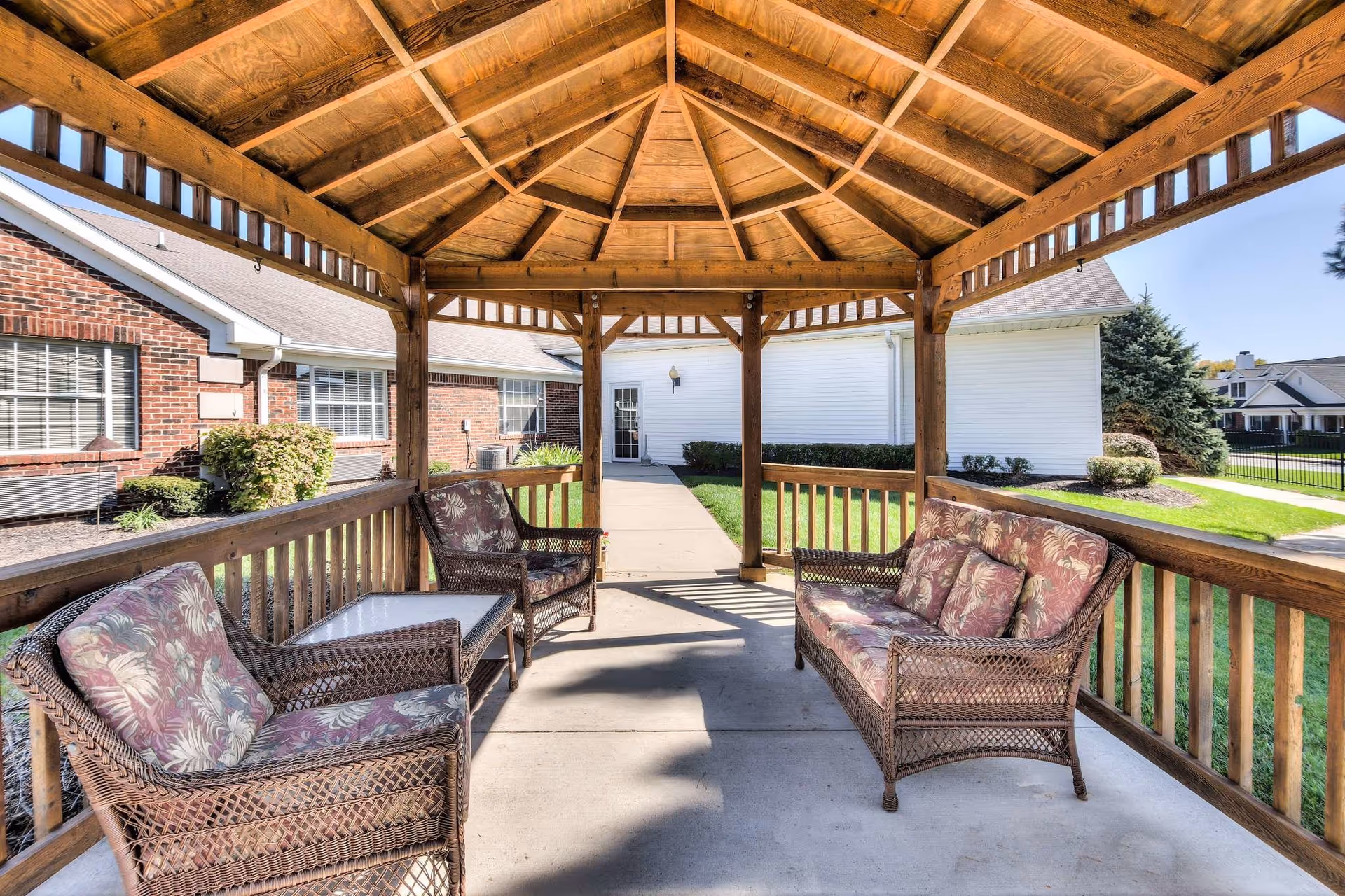 Wooden gazebo with wicker chairs and a cushioned sofa overlooking a courtyard between brick and white buildings.