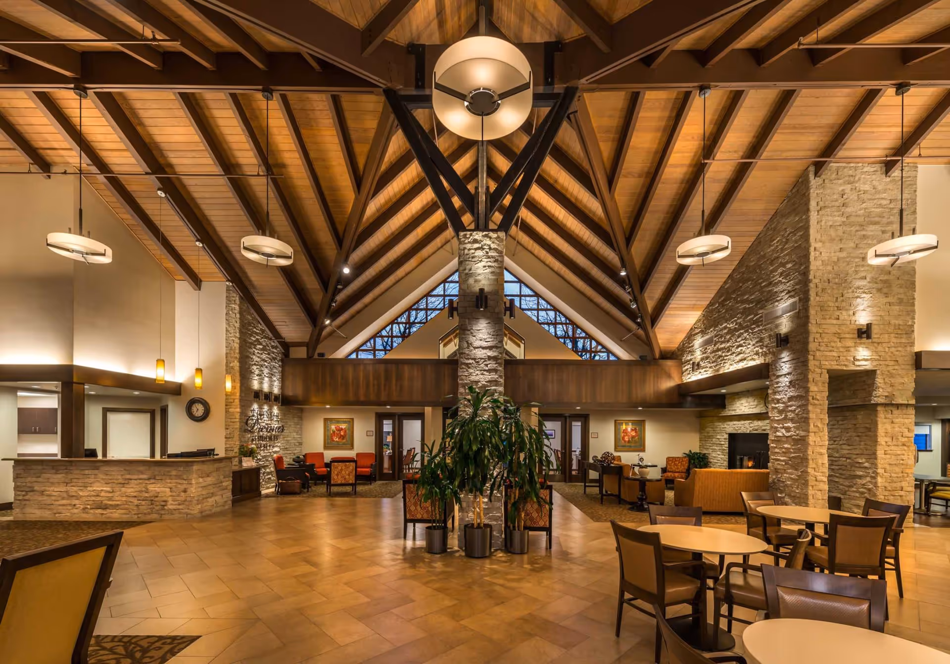 Spacious and warmly lit interior of a senior living facility with high vaulted wooden ceilings, stone pillars, and modern hanging light fixtures. The room features multiple seating areas with chairs and tables, a reception desk on the left, and a fireplace on the right. Large windows at the far end allow natural light to enter.
