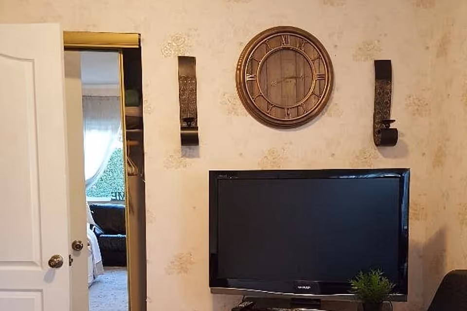Interior view of a living room area with a flat-screen TV mounted on a stand below a round wall clock with Roman numerals. The wall has a light floral wallpaper and two decorative wall sconces on either side of the clock. A partially open door reveals another room with a window covered by sheer curtains and a dark sofa.