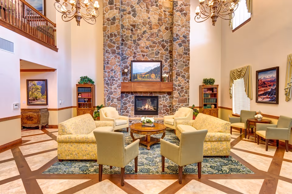 Comfortable communal living room with sofas and armchairs arranged around a central coffee table and a tall stone fireplace under chandeliers.