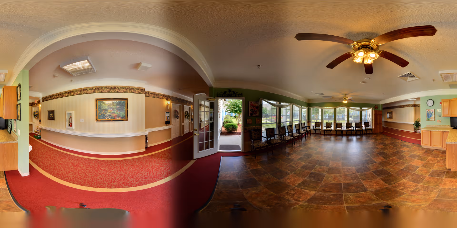 Panoramic view of an interior common area in a senior living facility with a red carpeted hallway on the left and a tiled floor seating area on the right. The seating area has multiple chairs lined up against large windows with a view of greenery outside. The ceiling has two ceiling fans with lights, and the walls are decorated with paintings and light fixtures. There is an open door leading outside in the center of the image.