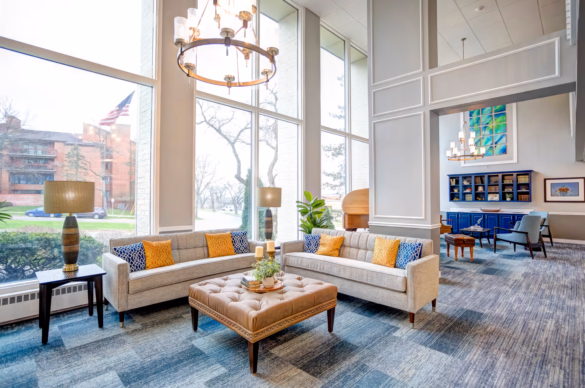 A bright and spacious living room area in The Gardens Retirement Community featuring two beige sofas with colorful blue and yellow patterned pillows, a large tufted ottoman in the center with decorative items on top, two side tables with lamps, large floor-to-ceiling windows letting in natural light, and a blue cabinet with shelves and artwork in the background.