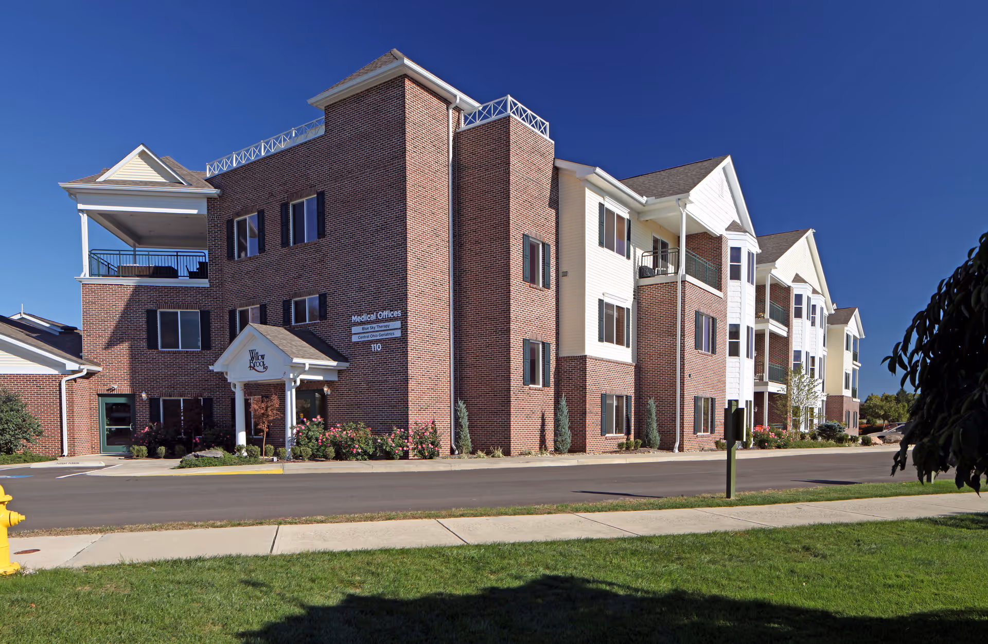 Exterior view of a multi-story senior living facility building with brick and white siding, balconies, and a clear blue sky. There is a sign indicating medical offices and a well-maintained lawn and sidewalk in front.
