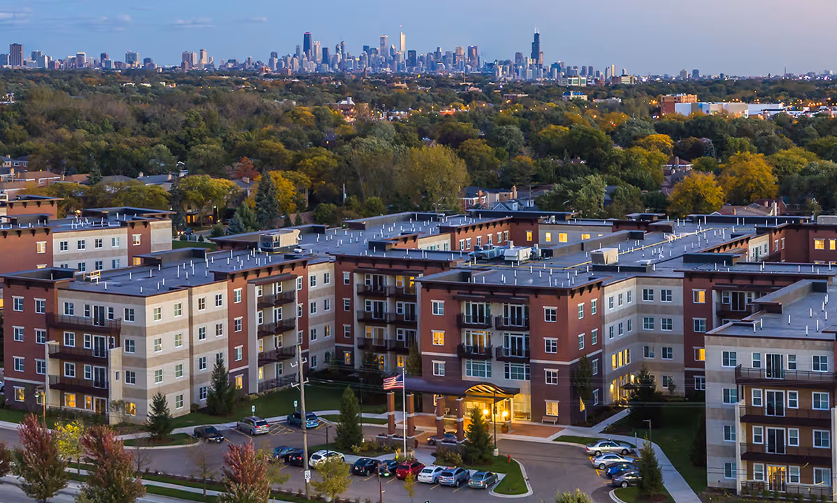 Aerial view of The Carrington at Lincolnwood senior living facility with multiple connected buildings surrounded by trees and parking lots, with a city skyline visible in the background during dusk.