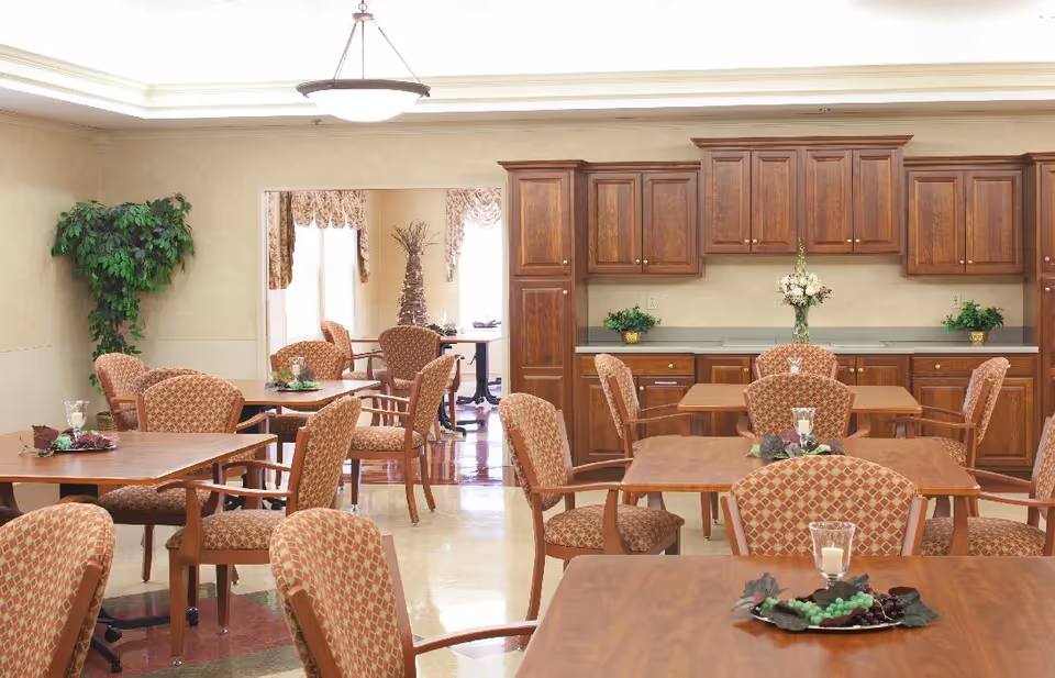 A dining room with multiple wooden tables and cushioned chairs arranged neatly. The room features wooden cabinets along one wall with decorative plants and a flower arrangement on the countertop. There is a large potted plant in the corner and a chandelier hanging from the ceiling. The room is well-lit with natural light coming from an adjacent room visible through an open doorway.