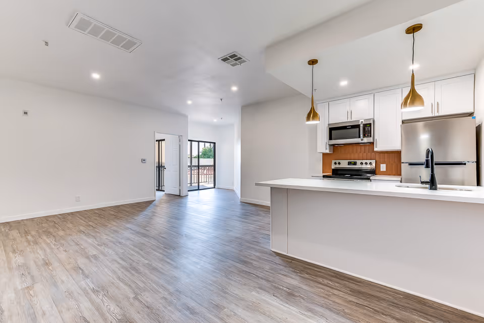 A modern open-concept interior space featuring a kitchen with white cabinets, stainless steel appliances, and two gold pendant lights hanging over a white countertop island. The room has light wood flooring and white walls, with a doorway leading to another room and a sliding glass door opening to a balcony or outdoor area.