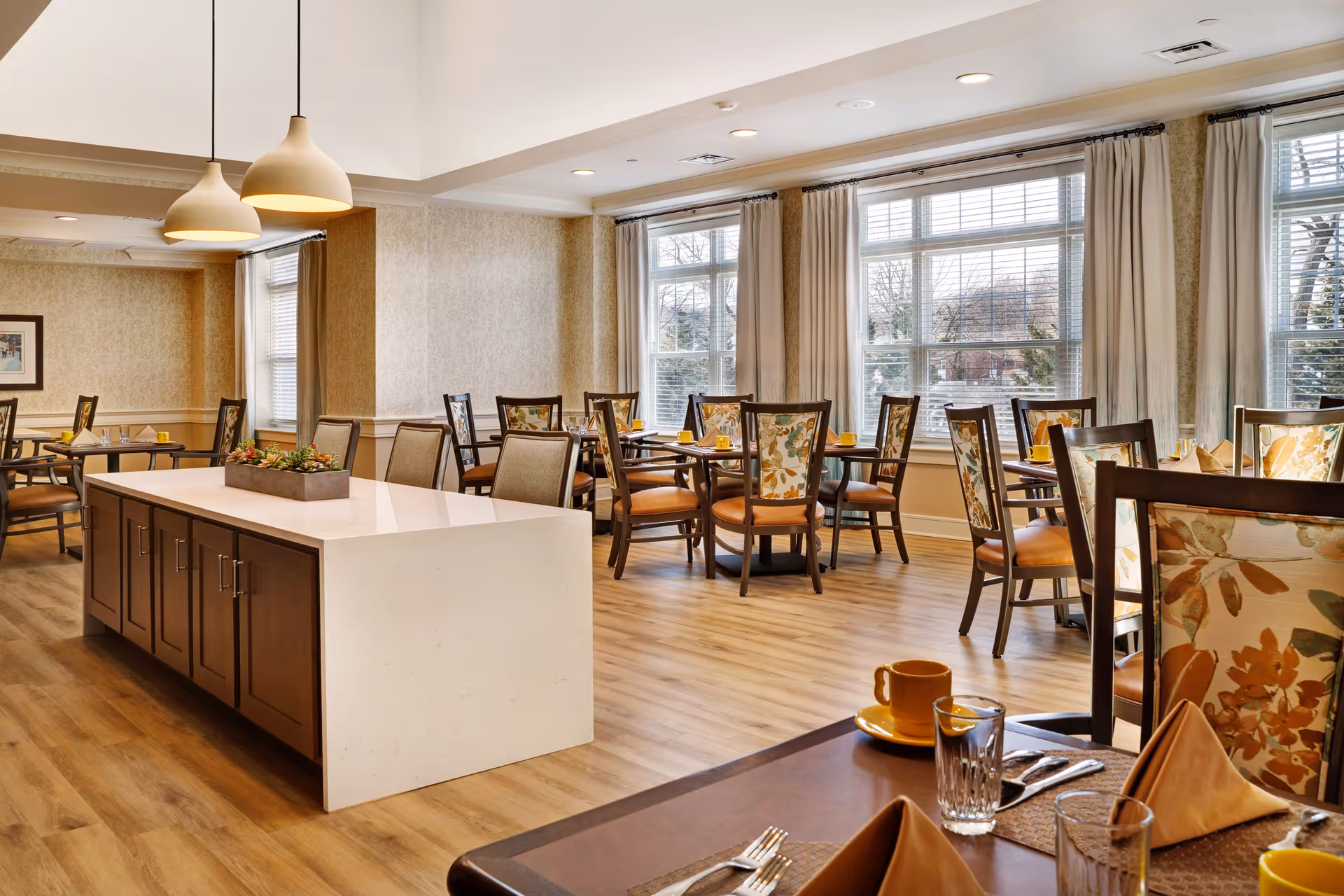 A bright and spacious dining room with multiple tables and chairs arranged neatly. The chairs have floral patterned backs and orange seats. Each table is set with yellow cups, glasses, and folded napkins. Large windows with white blinds and beige curtains allow natural light to fill the room. Two pendant lights hang above a central island with a white countertop and wooden cabinets below. The floor is light wood, and the walls have a subtle patterned wallpaper.