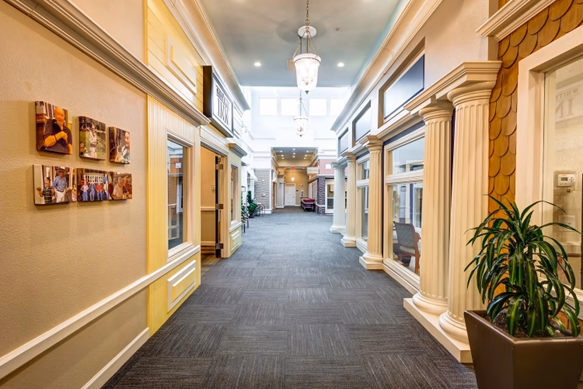 Well-lit interior hallway of a senior living facility with storefront-style rooms, decorative columns, carpeted floor, and hanging chandeliers.