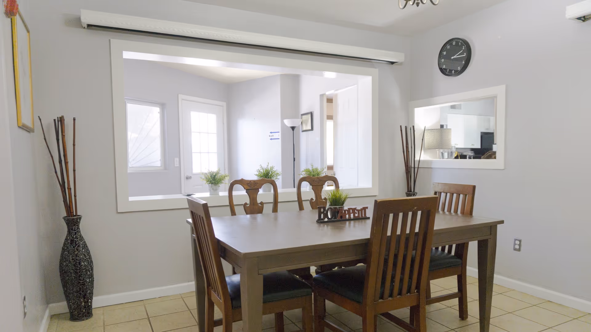 A dining room with a wooden table and four chairs. The table has a small decorative plant and a 'Bon Appetit' sign. The room has light gray walls, a large window opening to another room, a wall clock, a mirror, and a tall decorative vase with sticks in the corner.