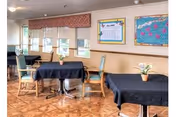 Interior view of a dining area with several tables covered in black tablecloths, each with a small potted plant centerpiece. Wooden chairs with cushions surround the tables. The room has large windows with blinds and valances, and colorful bulletin boards are displayed on the walls.