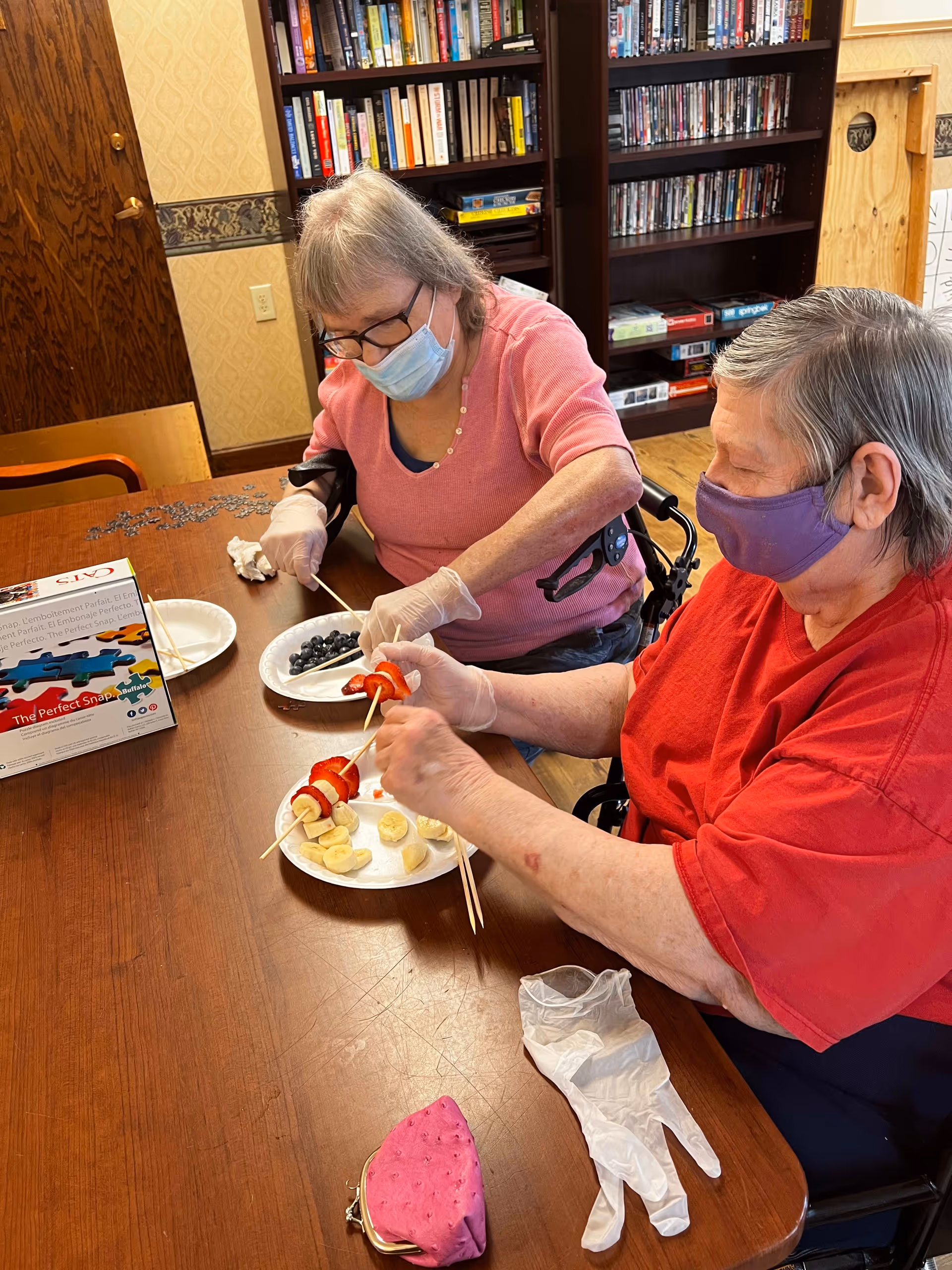 Two elderly women wearing face masks and gloves are sitting at a wooden table in a room with bookshelves. They are preparing fruit skewers using strawberries, bananas, and blueberries on paper plates. One woman is wearing a pink shirt and glasses, while the other is wearing a red shirt.
