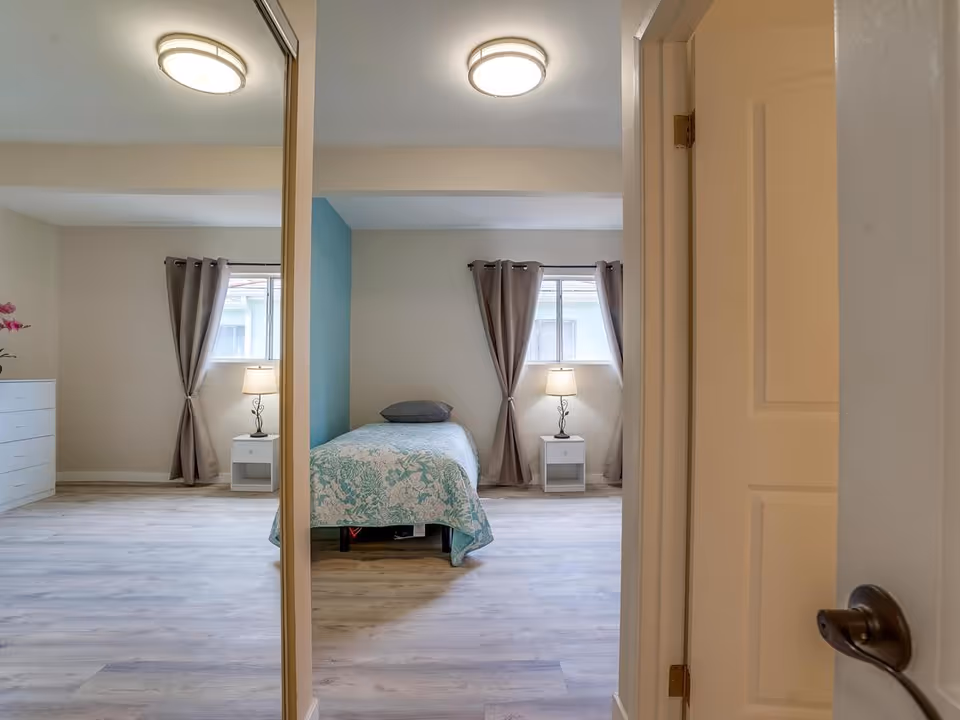 View through an open door into a bright single bedroom with a bed, nightstands and lamps, mirrored closet and curtains.