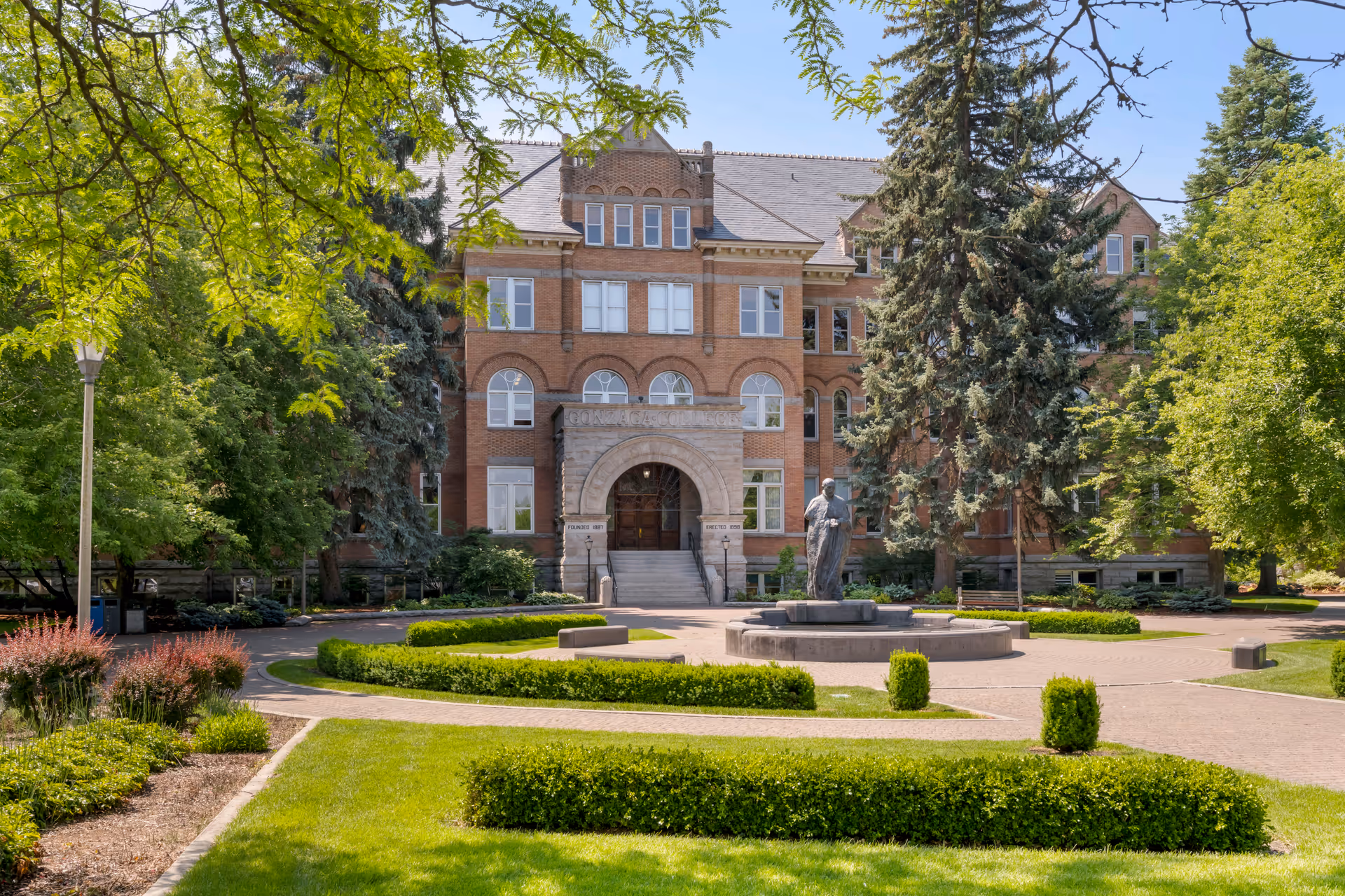 A large, historic brick building with multiple windows and an arched entrance, surrounded by well-maintained green lawns, bushes, trees, and a circular paved area with a statue in the center under a clear blue sky.