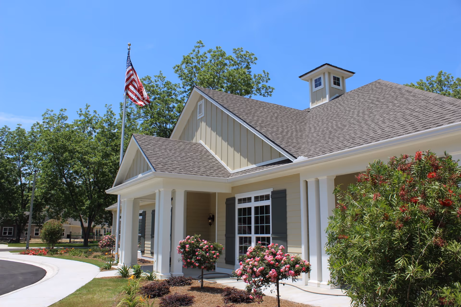 Exterior view of a single-story building with beige siding, white columns, and a gray shingled roof under a clear blue sky. There is an American flag on a flagpole in front of the building, surrounded by landscaped bushes and flowering plants.