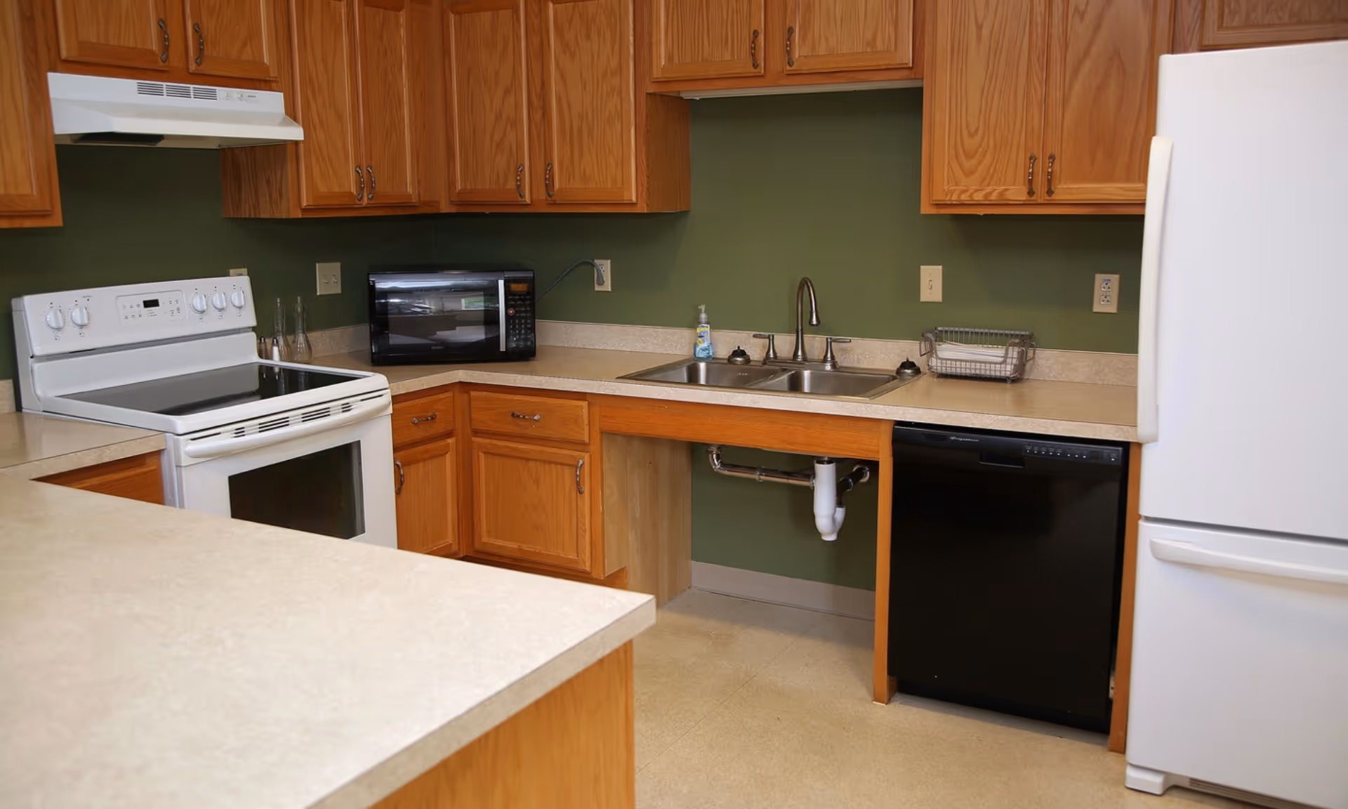Kitchen with oak cabinets, a white stove and refrigerator, microwave, double sink and black dishwasher against a green wall.