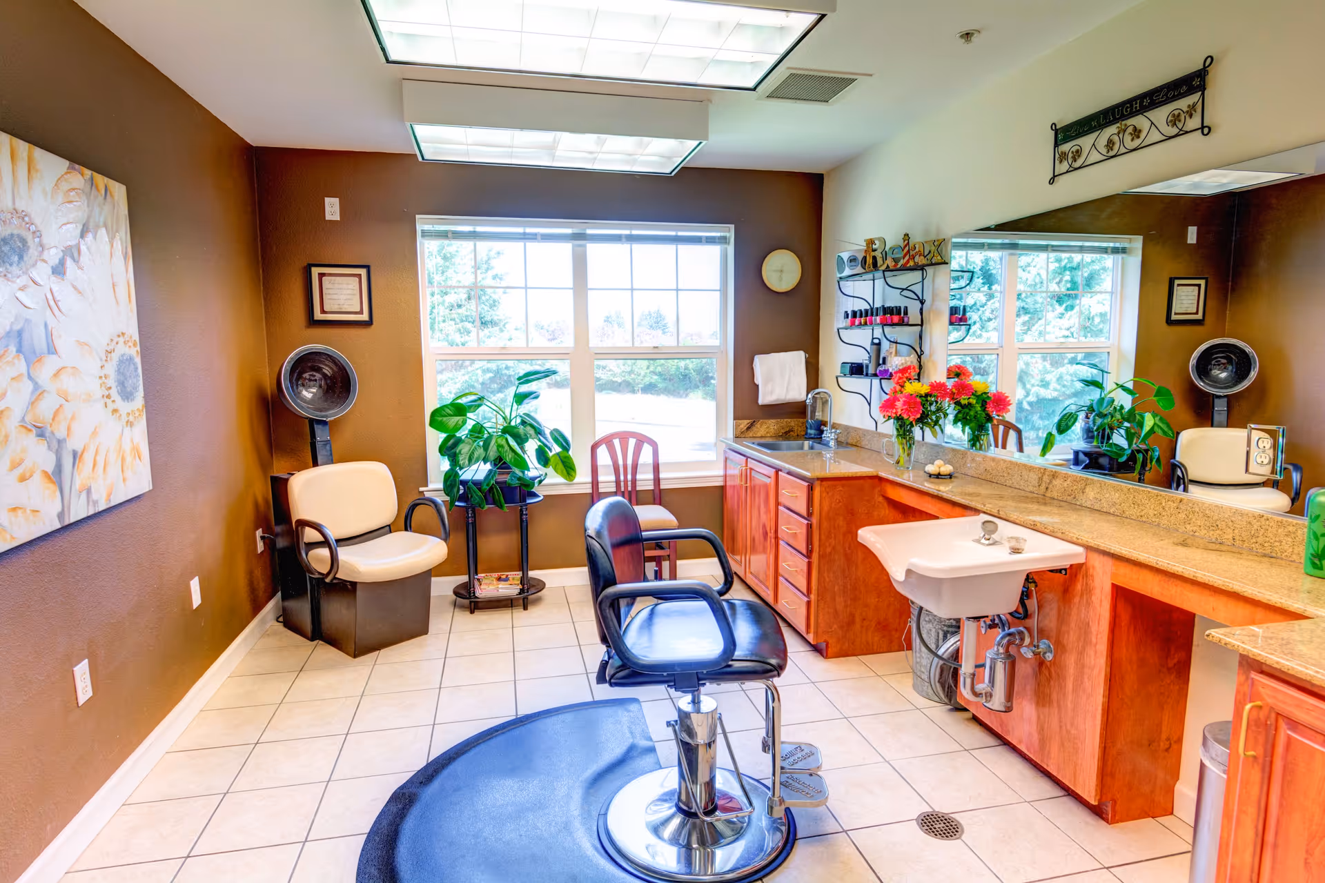 A bright and clean hair salon area inside an assisted living facility with a black salon chair on a black mat, a beige chair with a hair dryer, a large mirror above a wooden counter with a sink, plants, flowers, and nail polish racks, and a window letting in natural light.