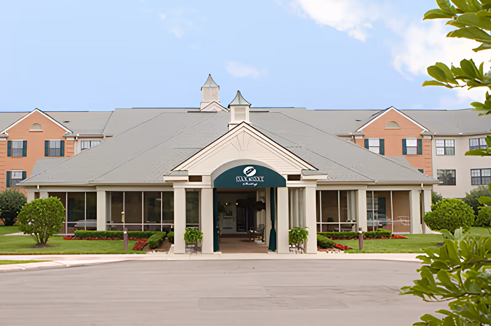 Front exterior view of Oakmont Livonia Senior Community building with a covered entrance, green awning, manicured bushes, and a clear blue sky.