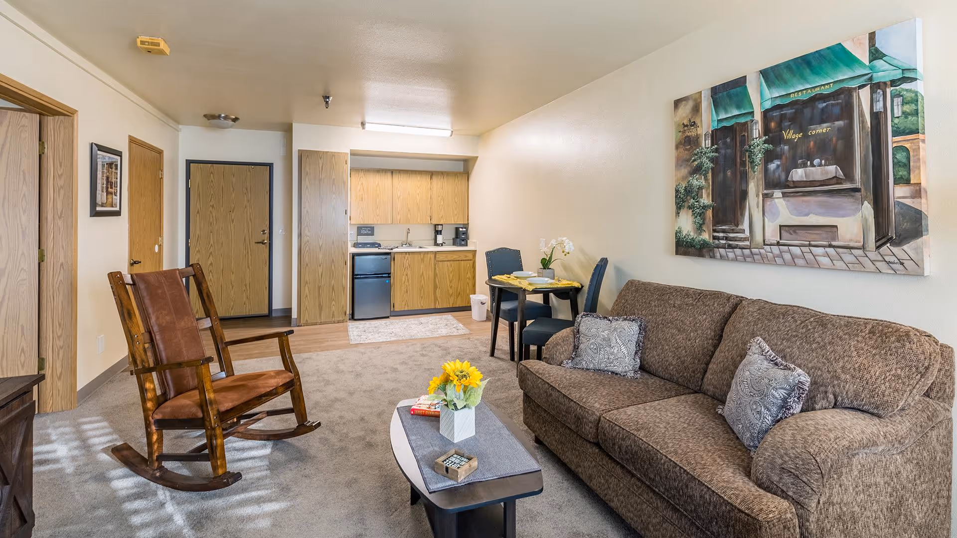 A cozy living room area in a senior living facility featuring a brown upholstered sofa with two decorative pillows, a wooden rocking chair with a brown cushion, a small coffee table with a vase of yellow flowers and books, and a small dining table with two chairs near a kitchenette with wooden cabinets, a mini fridge, and a coffee maker. A painting of a restaurant storefront hangs on the wall above the sofa.