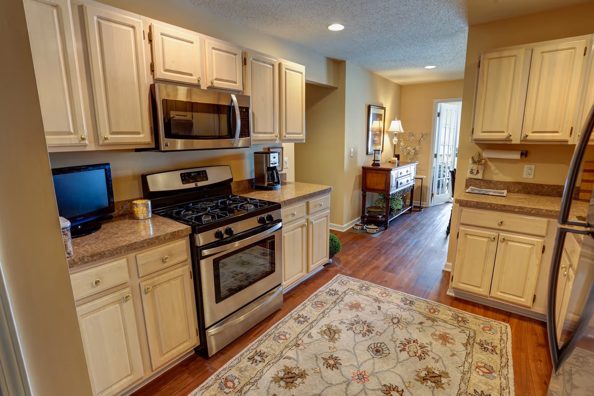 A modern kitchen featuring light wood cabinets, stainless steel appliances, and a decorative rug on the floor. The kitchen includes a gas stove, microwave, and a small TV on the countertop, with a glimpse of a dining area in the background.