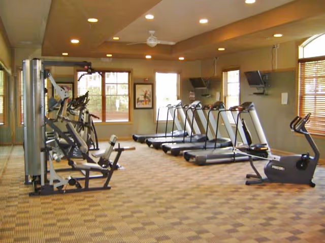 A fitness room with several exercise machines including treadmills, stationary bikes, and weight machines. The room has large windows with wooden blinds, beige walls, and a checkered carpet floor. Ceiling lights and a ceiling fan are visible.