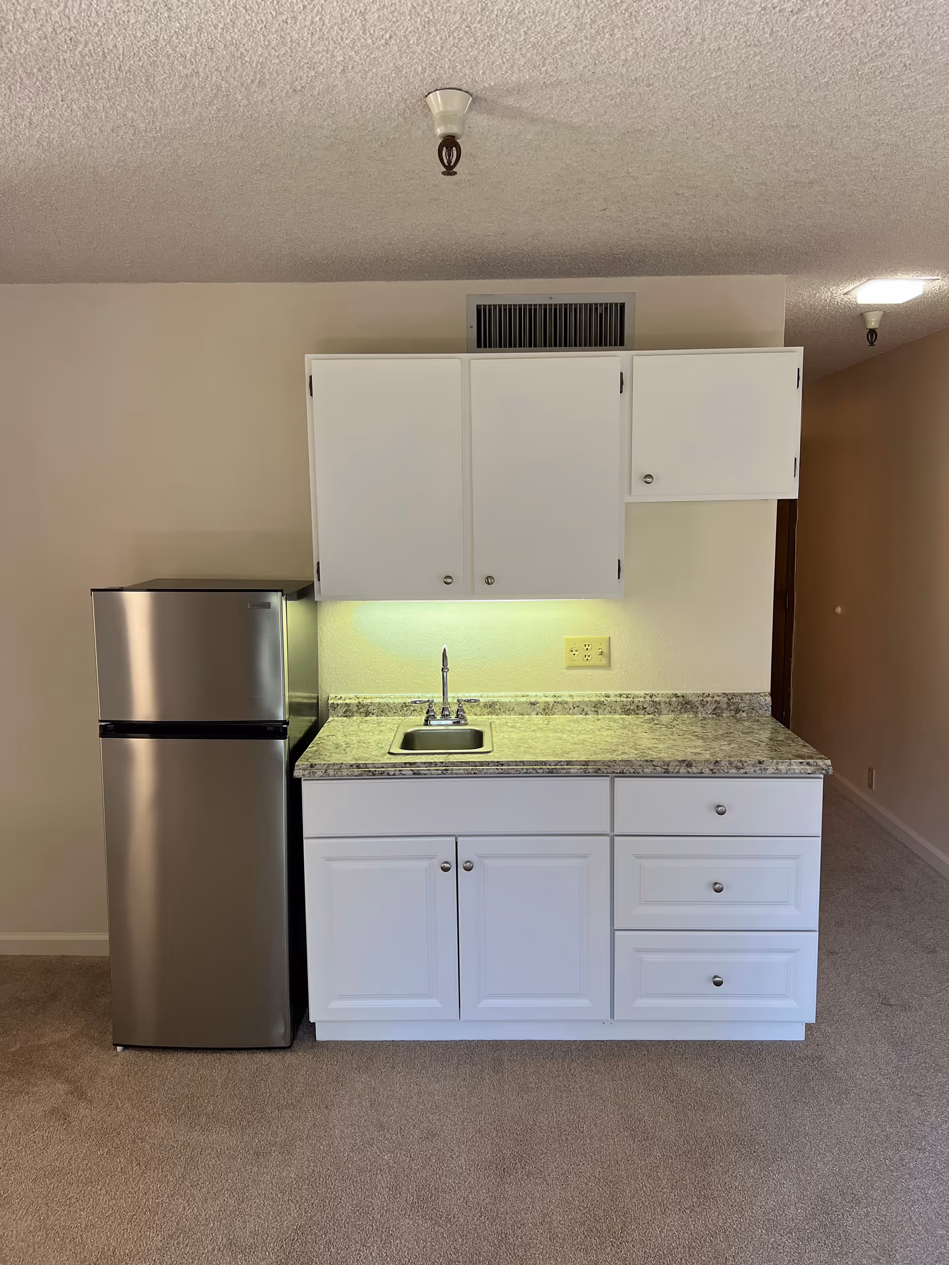 Compact kitchenette with a stainless steel refrigerator, white cabinets, marble-look countertop and a small sink in a carpeted room.