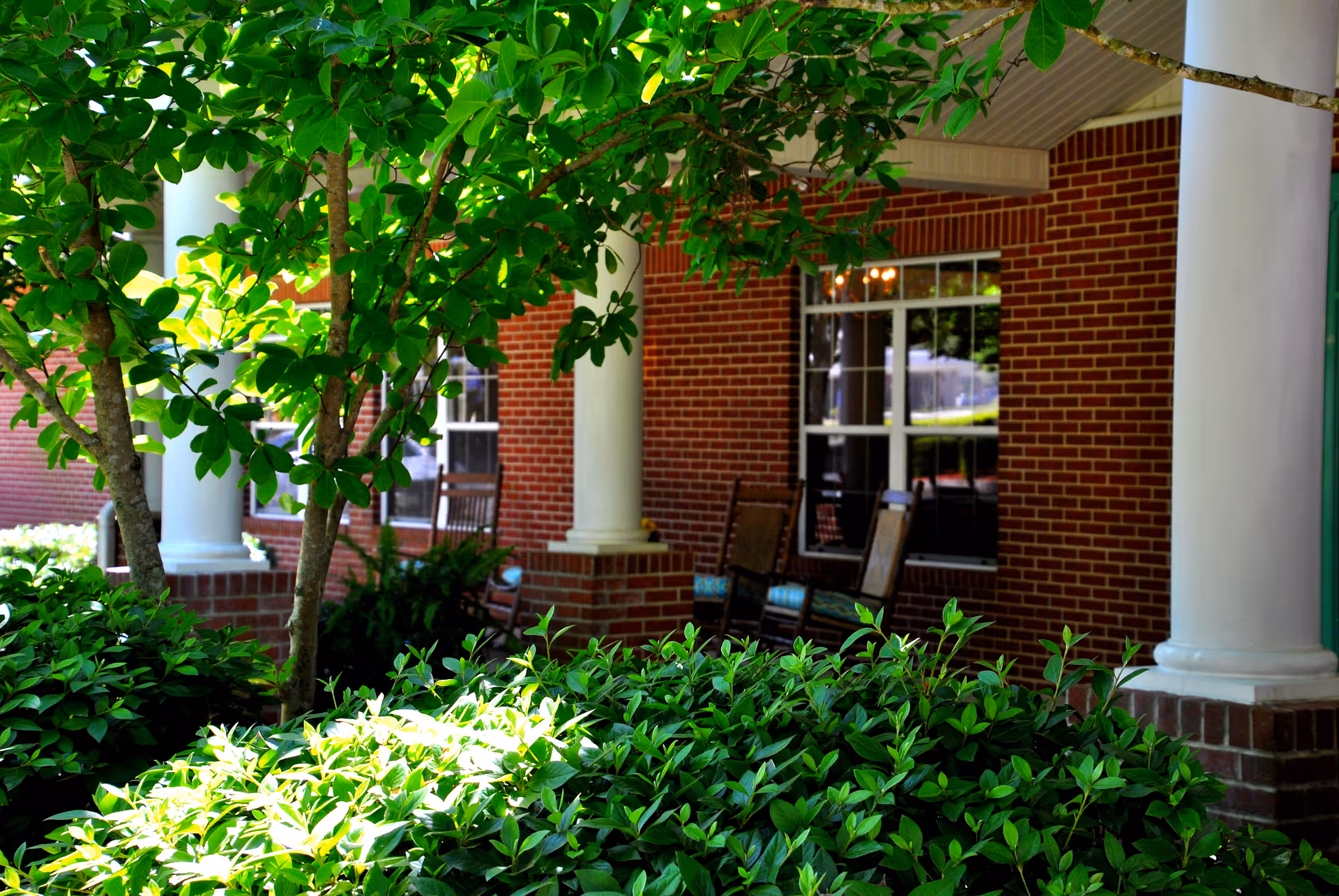 A shaded outdoor porch area with white columns and a brick wall. There are green leafy bushes and a tree in the foreground, and two wooden rocking chairs with cushions are visible near the windows on the porch.