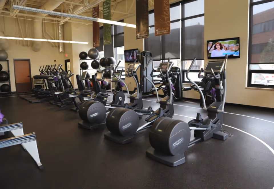 Bright fitness room with rows of elliptical machines, wall-mounted TVs, large windows, and stacked exercise balls.