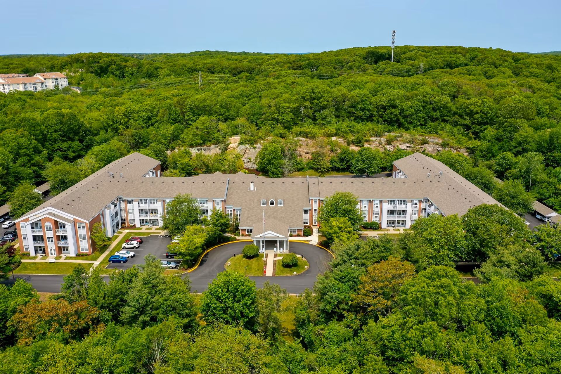 Aerial view of Solstice Senior Living at Groton, a large U-shaped senior living facility surrounded by dense green trees and vegetation, with a circular driveway and several parked cars in front.