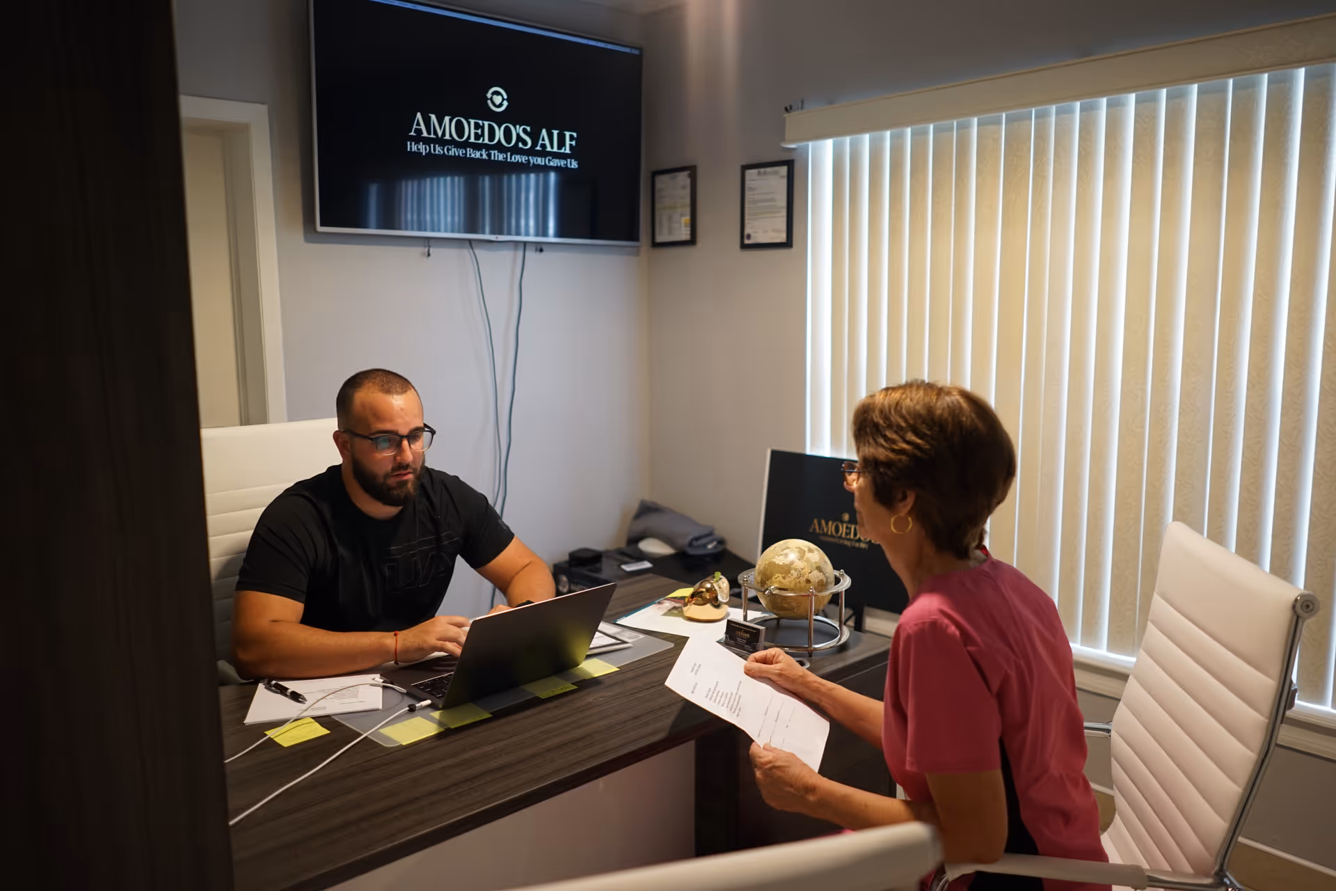 A man and a woman are seated across from each other at a desk in an office. The man is working on a laptop, and the woman is holding a document. Behind the man, a wall-mounted screen displays the text 'AMOEDO'S ALF Help Us Give Back The Love you Gave Us.' The office has vertical blinds covering the window and framed certificates on the wall.